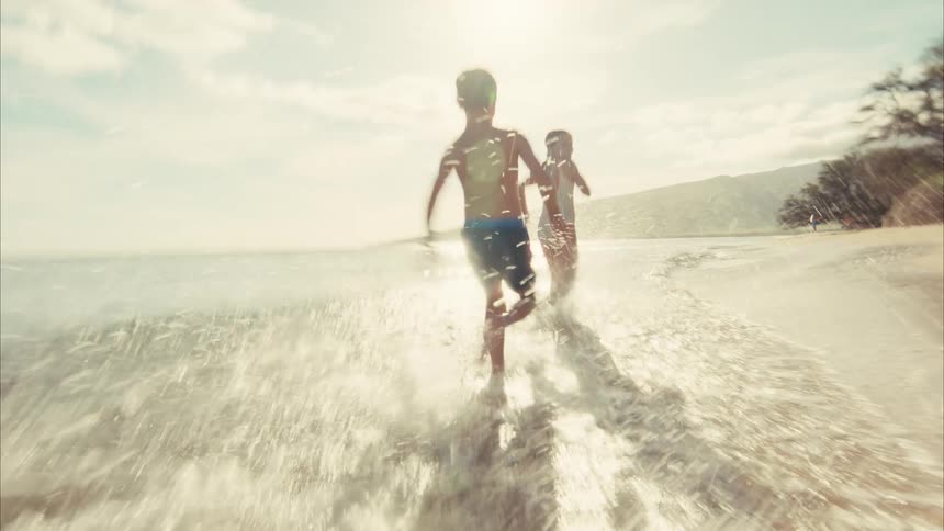 Slow-motion footage from a low angle shows two happy kids running and playing in the shallow waves of a sunny beach