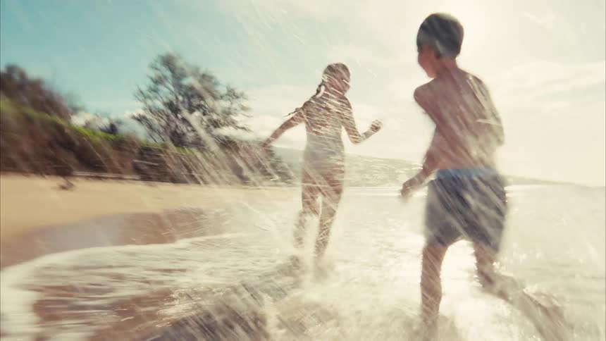 A boy and a girl playfully chase each other, running and splashing through the shallow ocean water along a sunny beach in slow motion