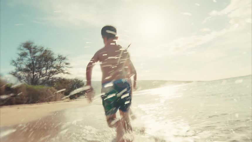 A boy and a girl playfully chase each other, running and splashing through the shallow ocean water along a sunny beach in slow motion