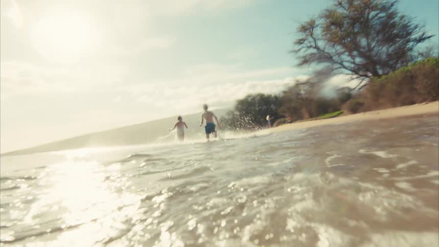 A happy young boy runs and splashes through the shallow ocean water in cinematic slow motion on a bright sunny day at the beach