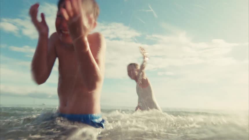 A boy and girl laugh and splash each other playfully in the ocean in a sun-drenched slow-motion shot from the water's surface