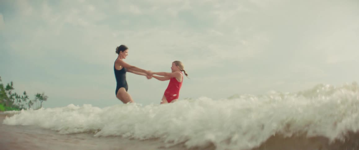A mother and daughter in swimsuits hold hands and playfully spin in the ocean waves in slow motion