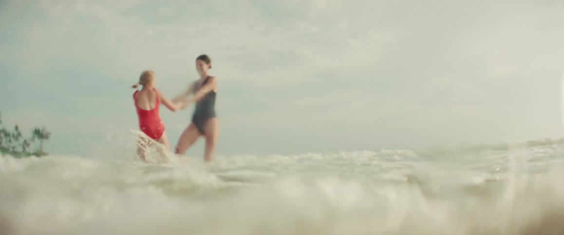 A mother and daughter in swimsuits hold hands and playfully spin in the ocean waves in slow motion