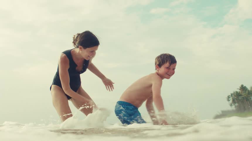 In an epic slow-motion shot, a mother and her son laugh and play as a wave splashes around them on a beautiful beach
