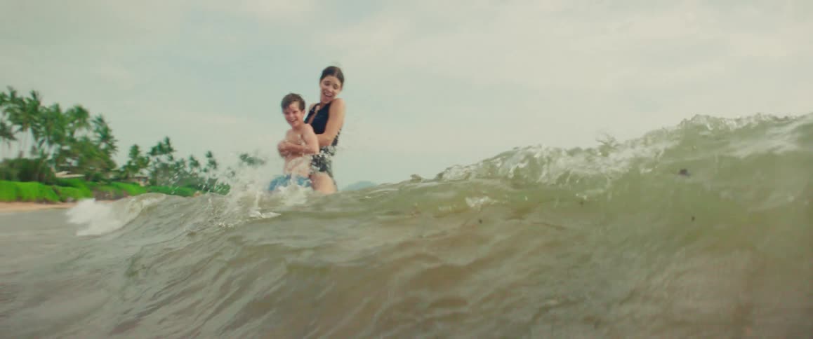 A happy mother holds her son as they brace for a wave to crash over them while playing in the ocean in slow motion