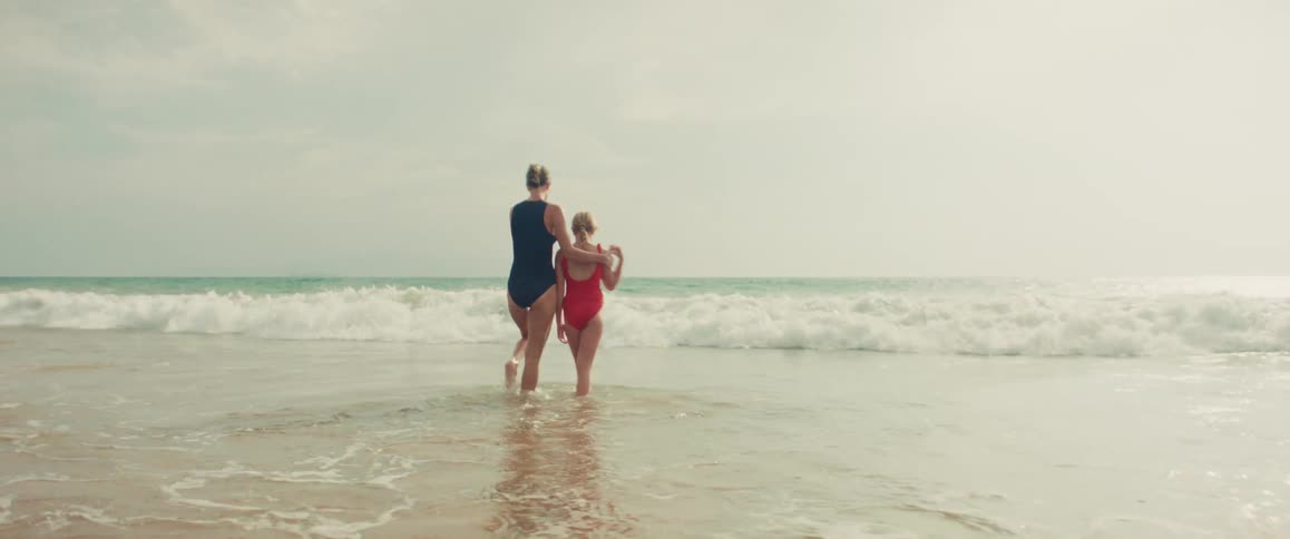 A mother and daughter share a loving embrace while standing in the ocean shallows, watching the waves roll in during a slow motion shot