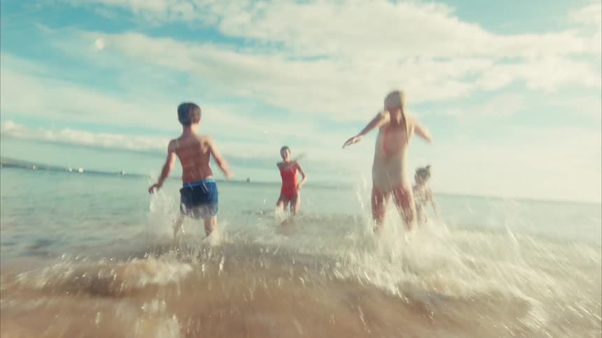 Joyful children run in slow motion across a sunny beach and splash into the shallow ocean waves