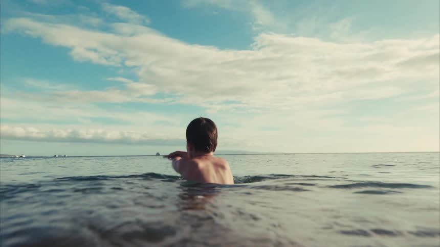 A young boy wades in the ocean before the camera submerges to follow him swimming underwater in slow motion
