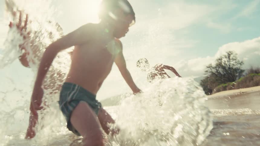 Joyful children run and splash through the ocean waves on a sunny tropical beach in slow motion