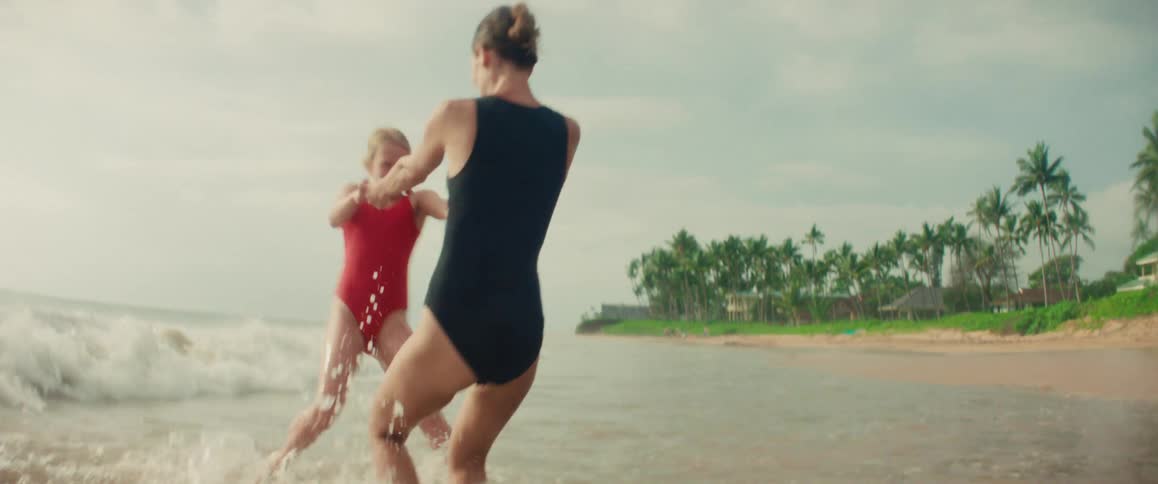 A happy mother and her young daughter playfully spin and splash in the shallow ocean waves on a tropical beach in slow motion