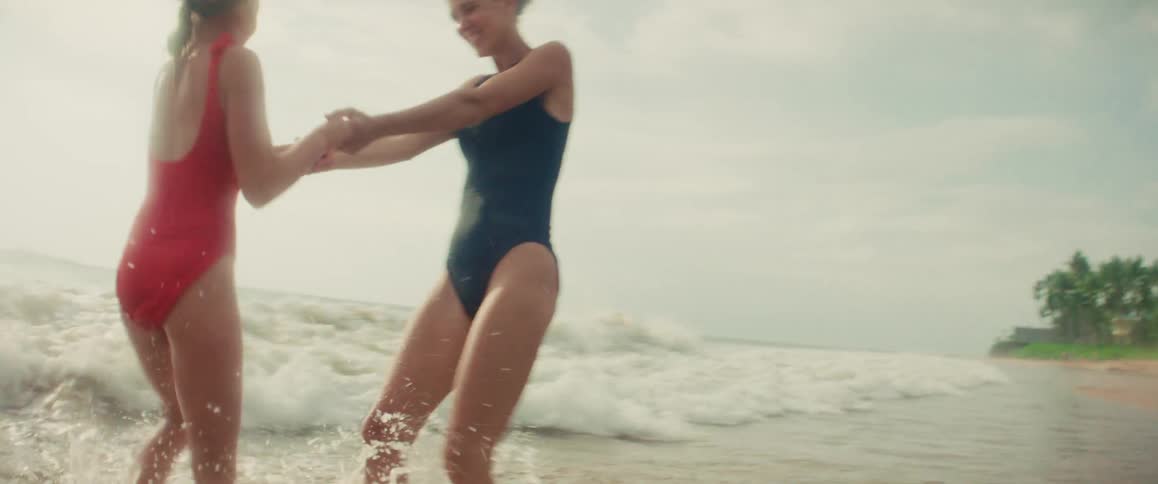 A happy mother and her young daughter playfully spin and splash in the shallow ocean waves on a tropical beach in slow motion