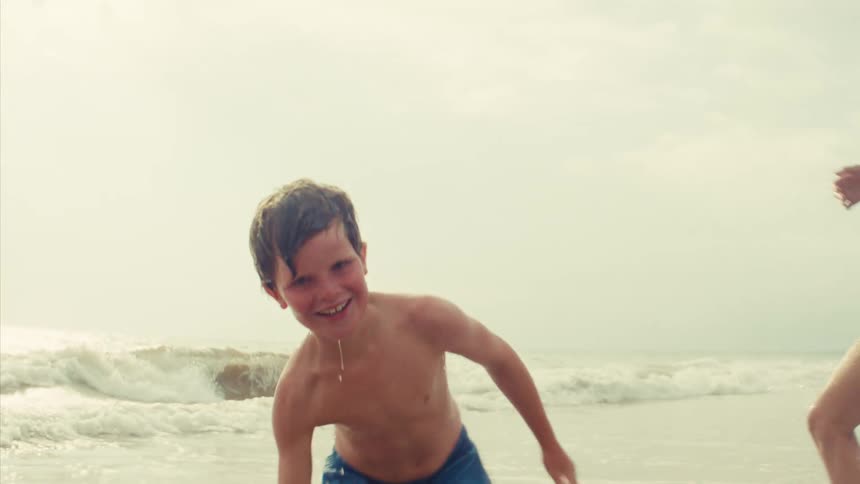 A happy young boy splashes water towards the camera in slow motion while playing with his mother in the ocean waves