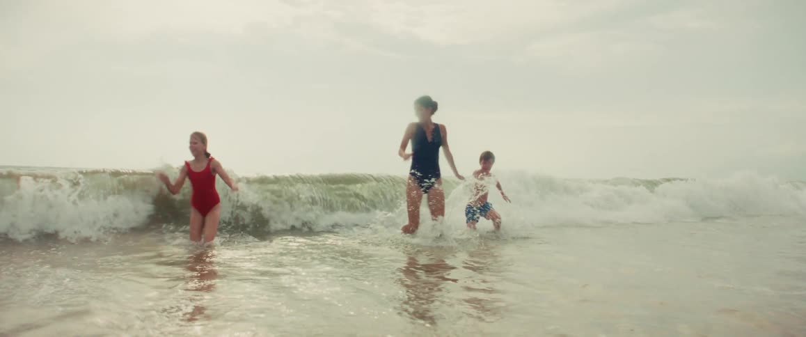 A mother and her two children laugh and play as a gentle ocean wave breaks around them in slow motion