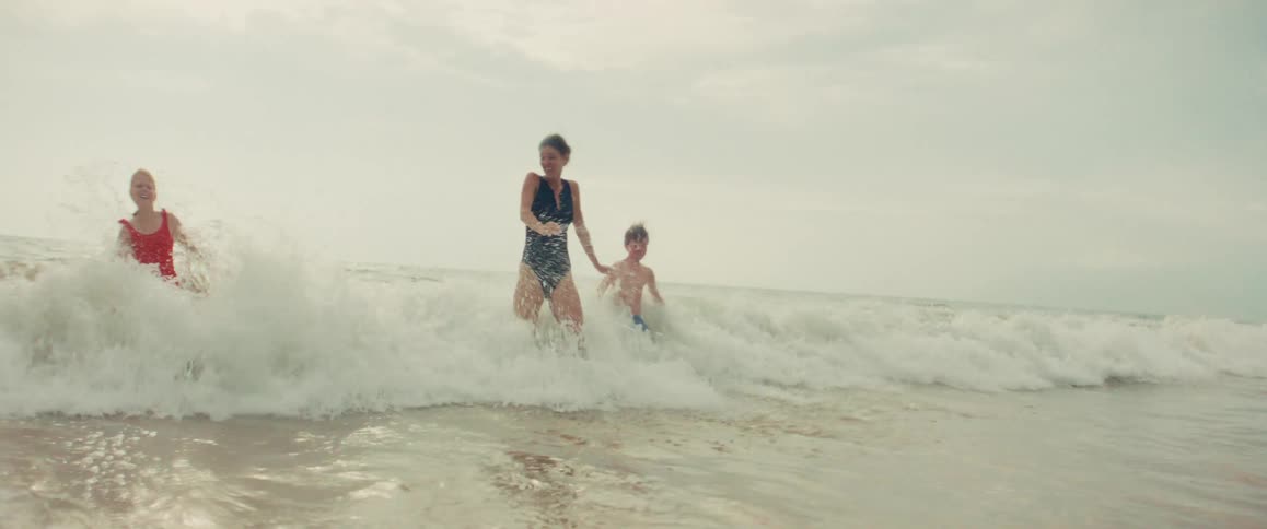 A mother and her two children laugh and play as a gentle ocean wave breaks around them in slow motion
