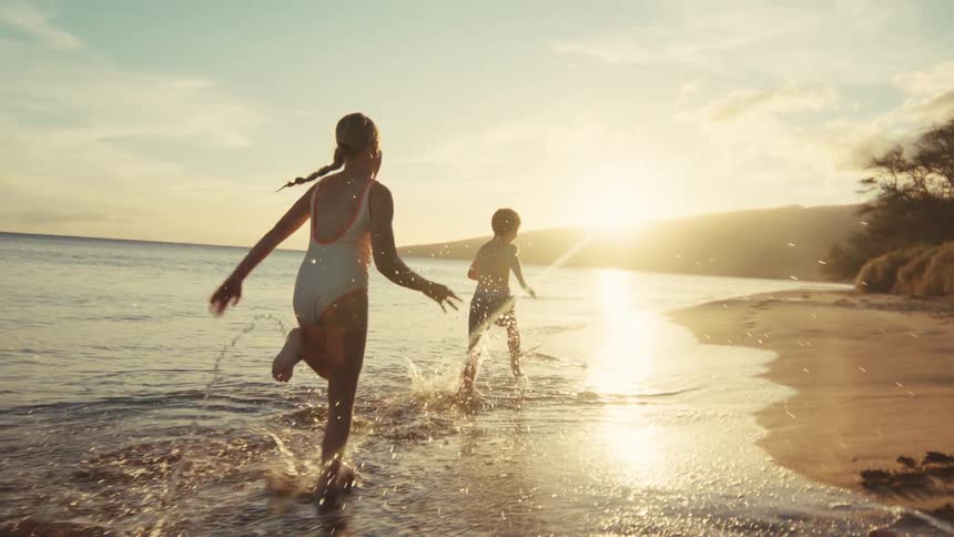 Two happy young children run and splash along the beach shoreline in the ocean at sunset in slow motion