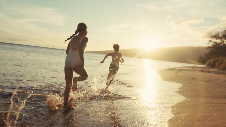 Two happy young children run and splash along the beach shoreline in the ocean at sunset in slow motion
