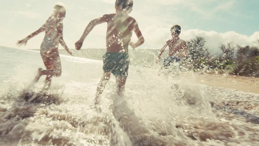 Three carefree children run and splash in the shallow ocean waves on a bright sunny day in slow motion