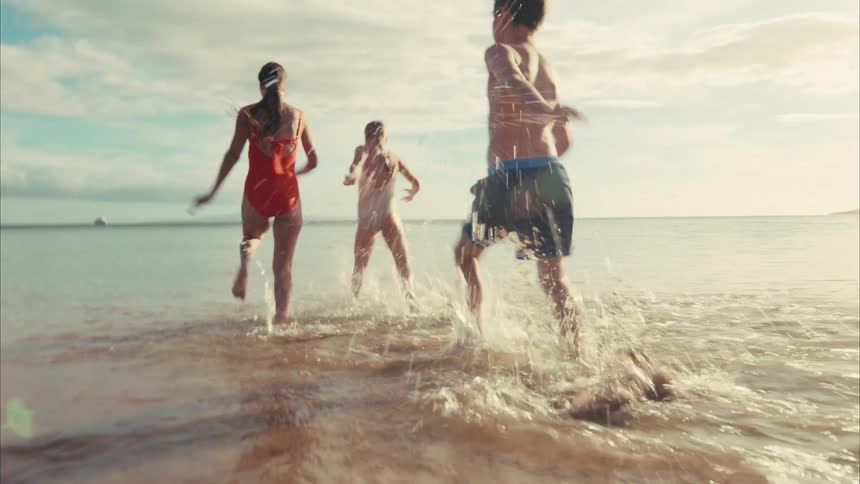 A mother and her two children laugh and run into the ocean in slow motion, splashing in the warm golden sunlight