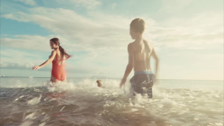 A mother and her two children laugh and run into the ocean in slow motion, splashing in the warm golden sunlight