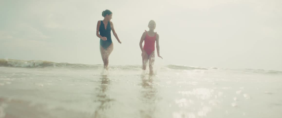 A mother and her young daughter happily run through the shallow ocean waves towards the camera in slow motion