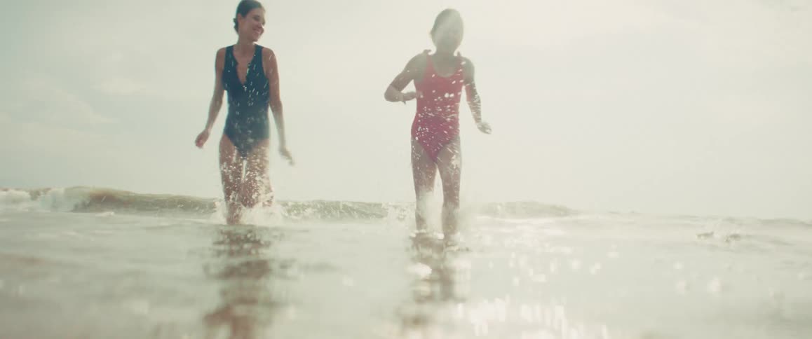 A mother and her young daughter happily run through the shallow ocean waves towards the camera in slow motion