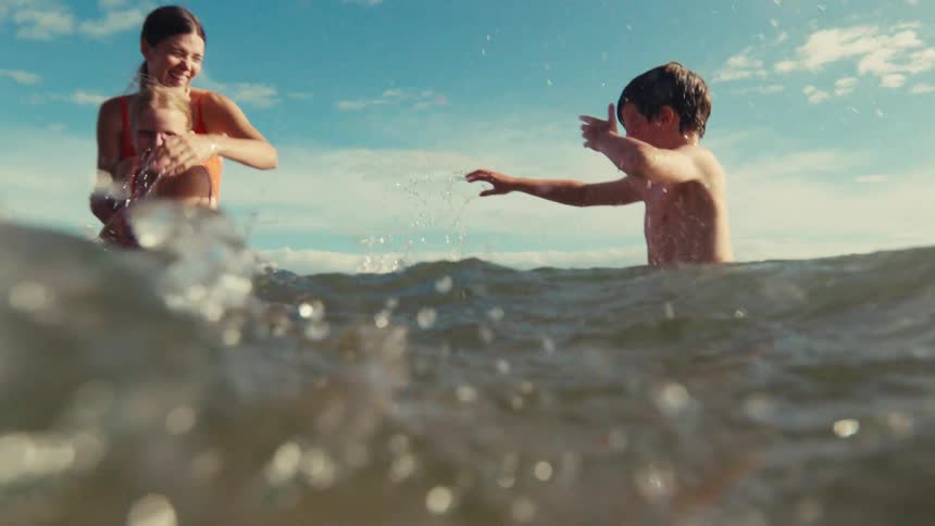 A happy mother plays with her son and daughter in the ocean, splashing water at each other in slow motion on a beautiful sunny day