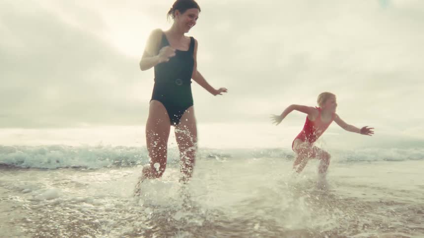 A mother and daughter joyfully run out of the ocean waves onto the beach in slow motion