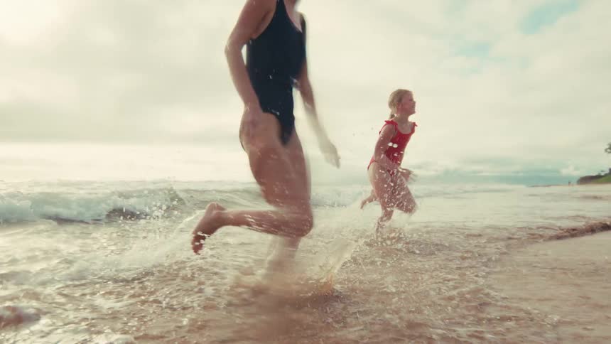 A mother and daughter joyfully run out of the ocean waves onto the beach in slow motion