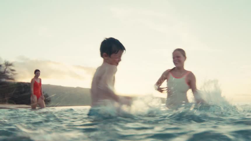 A young boy and girl laugh and joyfully splash each other while playing in the ocean at sunset in slow motion