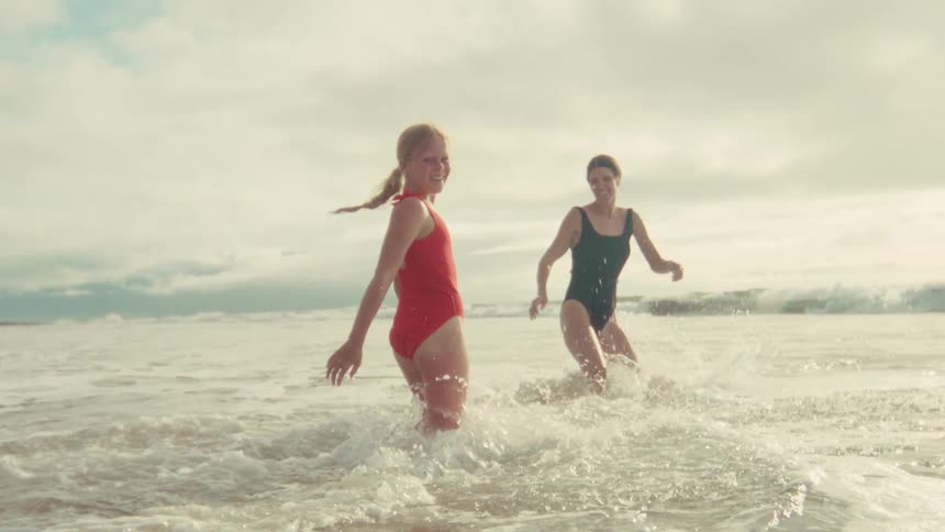 A smiling young girl in a red swimsuit looks back at the camera while wading in the ocean with her mother in slow motion