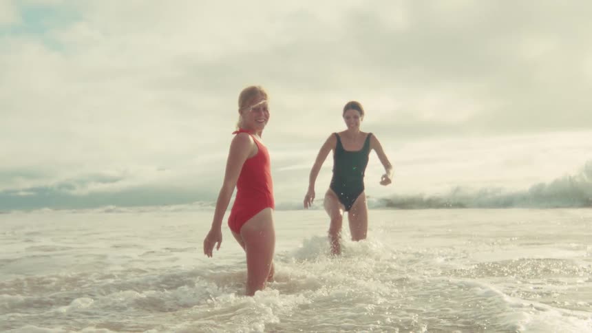 A smiling young girl in a red swimsuit looks back at the camera while wading in the ocean with her mother in slow motion