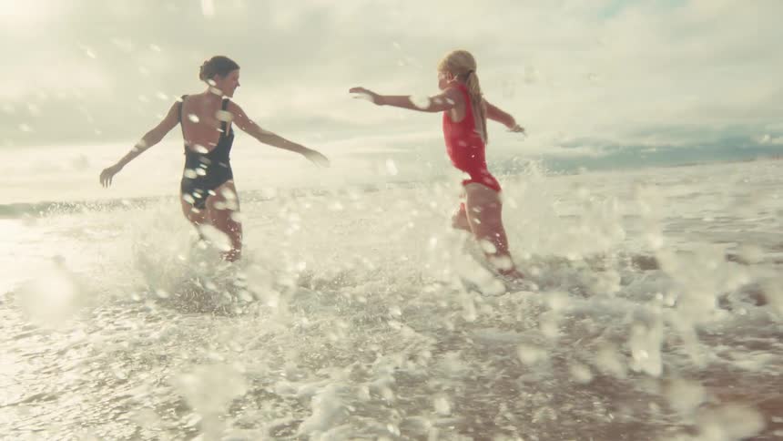 Two women in vintage-style swimsuits playfully splash and dance in the sunlit ocean surf in a beautiful slow-motion shot