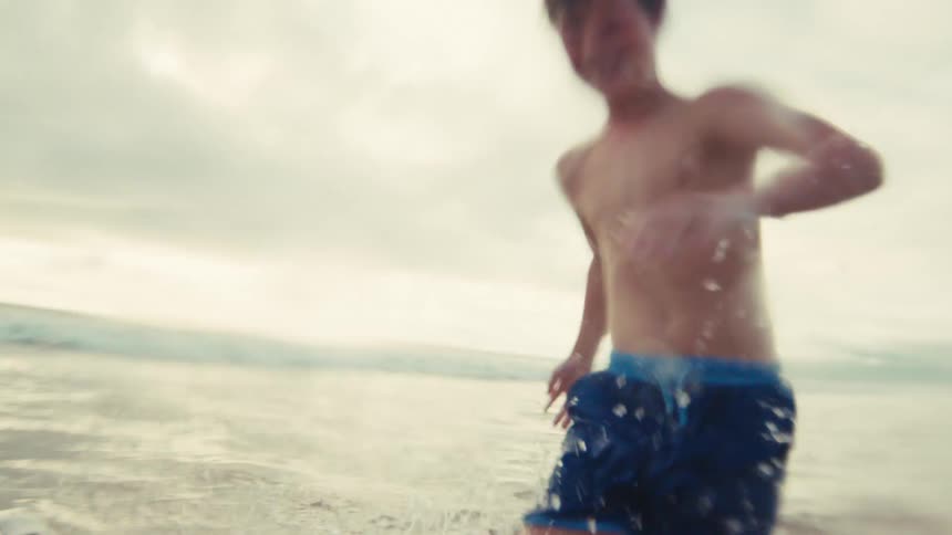 Joyful young boy laughs as he splashes water towards the camera while running in the ocean in slow motion