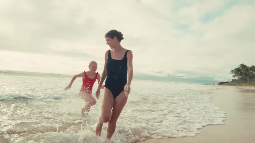 A happy mother and her daughter playfully run through the shallow ocean waves on a sandy beach in slow motion