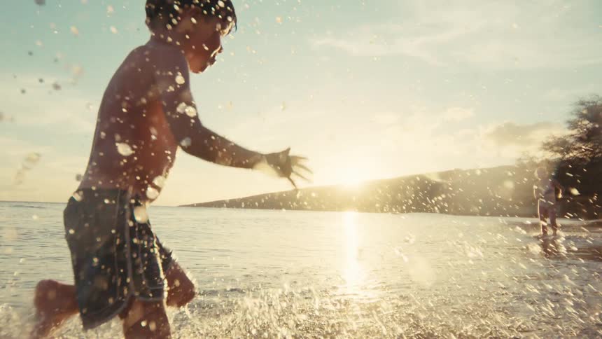 A joyful young boy splashes through the shallow ocean water as the sun sets in a beautiful, warm glow