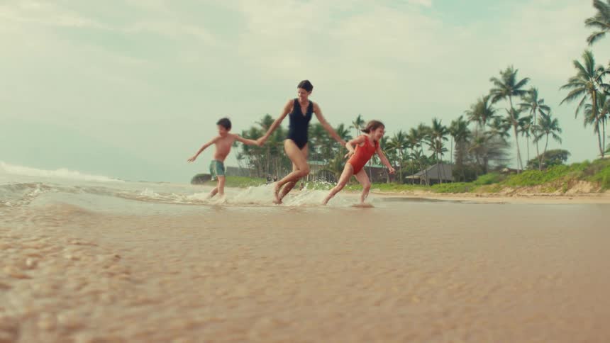 A mother and her two young children happily run and splash in the shallow water of a tropical beach in slow motion