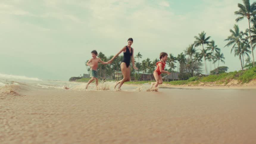A mother and her two young children happily run and splash in the shallow water of a tropical beach in slow motion