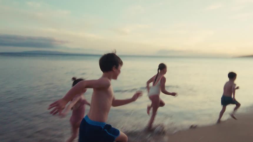 A group of energetic children playfully run in slow motion along a sandy beach shoreline at golden hour