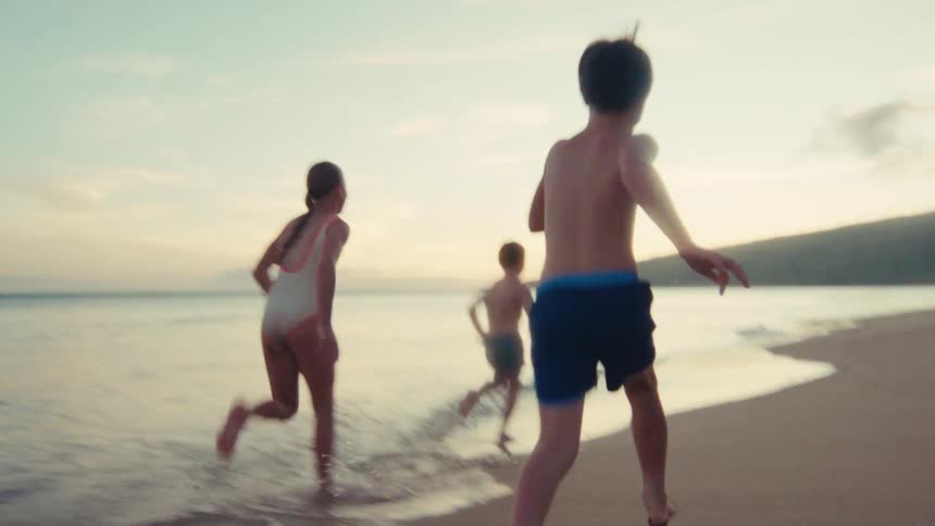A group of energetic children playfully run in slow motion along a sandy beach shoreline at golden hour