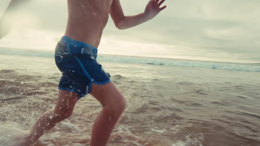 Close-up slow motion footage captures a young boy energetically running through the shallow ocean waves at the beach