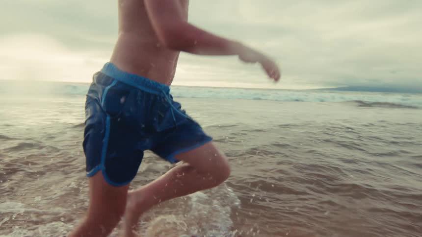Close-up slow motion footage captures a young boy energetically running through the shallow ocean waves at the beach
