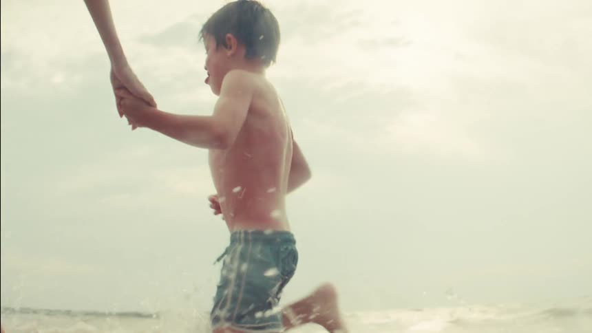 A mother and her young son run hand-in-hand, splashing through the ocean waves in slow motion