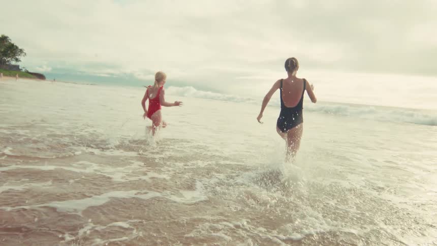 A mother and her young daughter happily run and play in the shallow ocean waves on a bright day in slow motion
