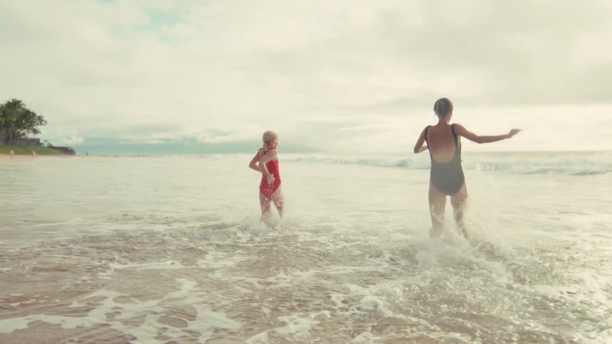A mother and her young daughter happily run and play in the shallow ocean waves on a bright day in slow motion