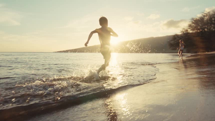 Slow motion shot of a young boy running and splashing along the ocean shore during a beautiful golden sunset