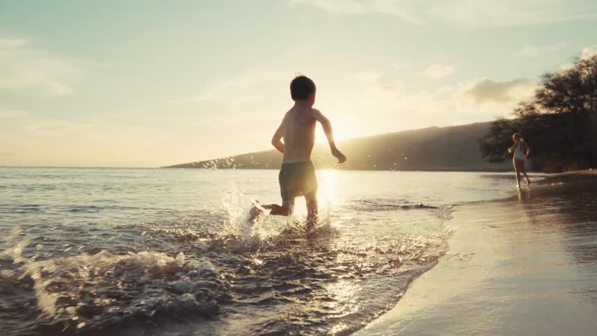 Slow motion shot of a young boy running and splashing along the ocean shore during a beautiful golden sunset