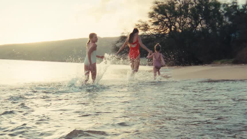 A mother holds hands with her two young daughters as they joyfully run and splash in the shallow ocean water at sunset in slow motion
