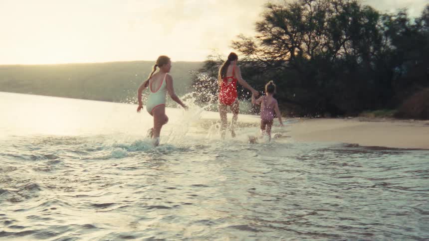 A mother holds hands with her two young daughters as they joyfully run and splash in the shallow ocean water at sunset in slow motion