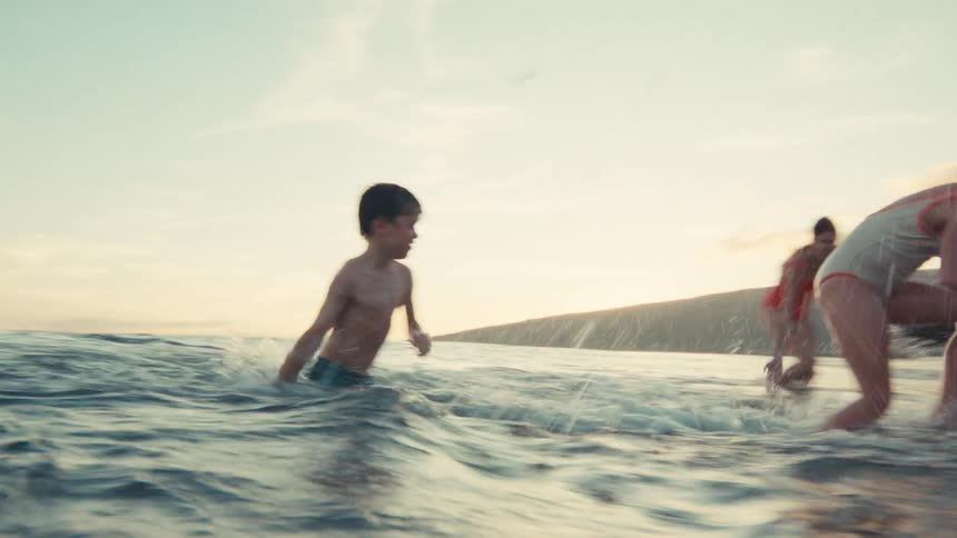 A joyful family with a young boy and girl splash each other in the ocean as they run towards the beach at sunset in slow motion