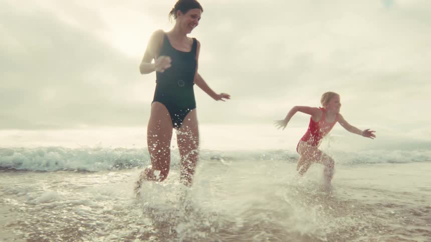 A happy mother and her young daughter joyfully run and splash through the shallow ocean waves together in slow motion